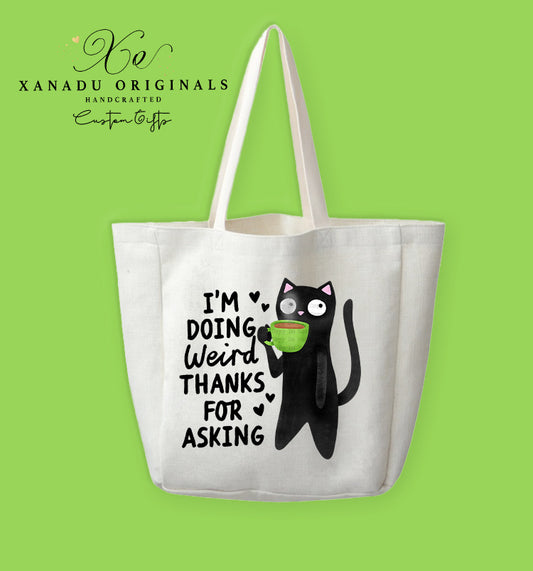 A cream-colored polyester linen tote bag featuring a cartoon illustration of a wide-eyed black cat holding a green mug filled with a pink warm drink. Bold black text to the left reads "I'M DOING Weird THANKS FOR ASKING" accented with tiny black hearts.