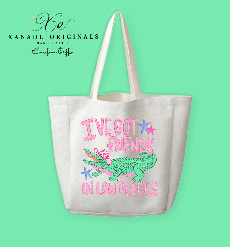 A cream-colored polyester linen tote bag featuring a cartoon illustration of a green alligator wearing a pink cowboy hat and a pink bandana, surrounded by stylized pink text reading "I'VE GOT FRIENDS IN LOW PLACES" accented with pink and blue star graphics.