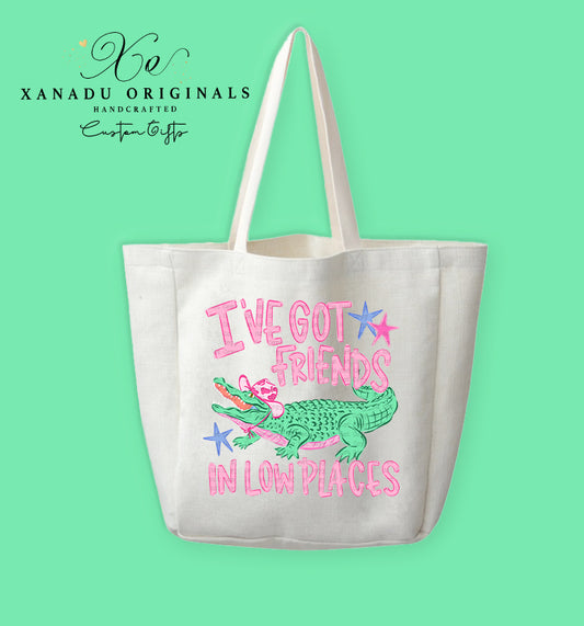 A cream-colored polyester linen tote bag featuring a cartoon illustration of a green alligator wearing a pink cowboy hat and a pink bandana, surrounded by stylized pink text reading "I'VE GOT FRIENDS IN LOW PLACES" accented with pink and blue star graphics.