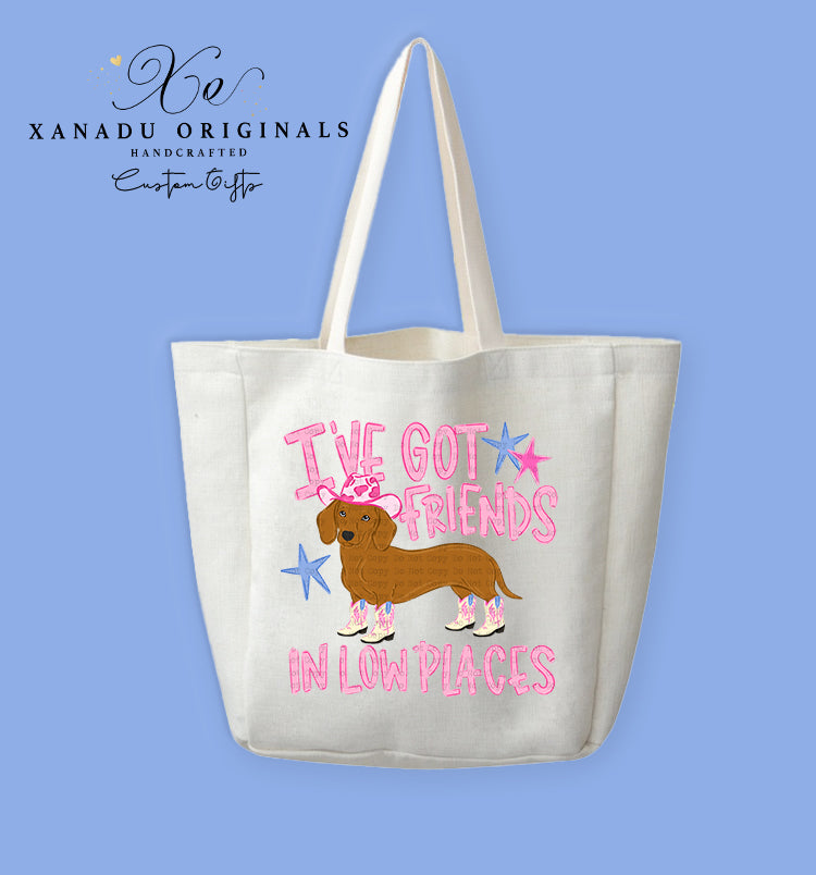 A cream-colored polyester linen tote bag featuring a cartoon illustration of a brown dachshund wearing a pink cowboy hat and four pink cowboy boots, surrounded by stylized pink text reading "I'VE GOT FRIENDS IN LOW PLACES" accented with pink and blue star graphics.