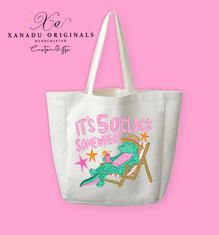 A cream-colored polyester linen tote bag featuring a cartoon illustration of a green alligator wearing a straw hat and drinking a pink cocktail while relaxing on a pink striped beach chair, under bold pink and yellow text reading "IT'S 5 O'CLOCK SOMEWHERE" with orange starfish.