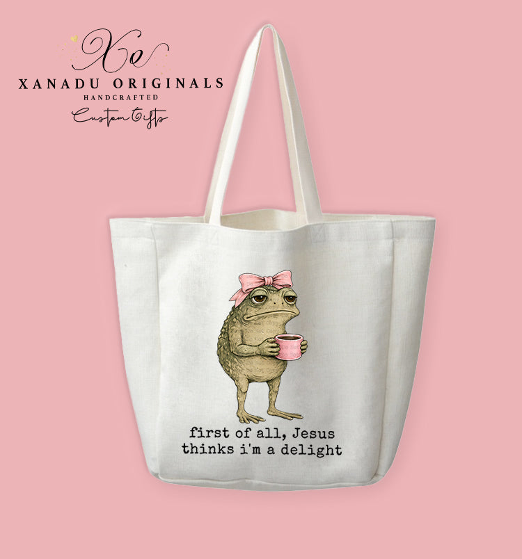 A cream-colored polyester linen tote bag featuring a watercolor illustration of a grumpy brown toad wearing a pink bow and holding a pink coffee cup, above black typewriter font text that reads "first of all, Jesus thinks i'm a delight."