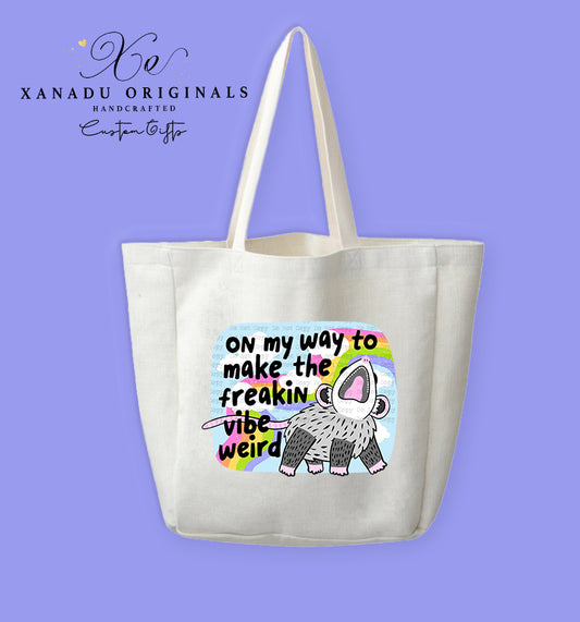 A cream-colored polyester linen tote bag featuring a cartoon illustration of a wide-eyed, screaming grey opossum with an open pink mouth. The animal is set against a bright rainbow backdrop with bold black text that reads "ON my way to make the freakin vibe weird."