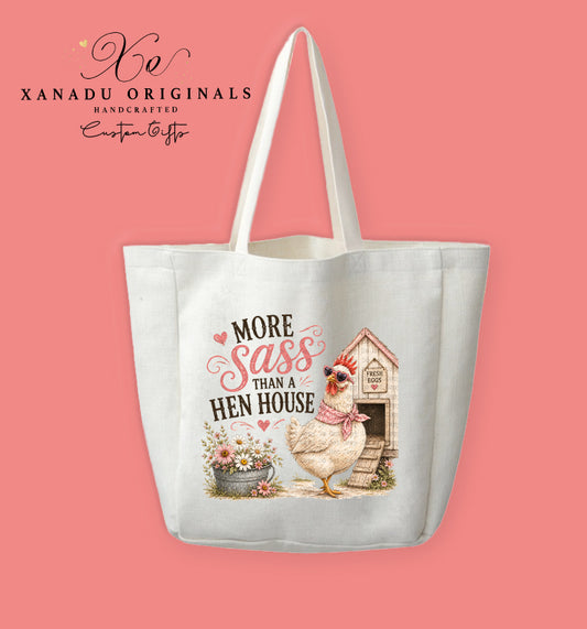 A cream-colored polyester linen tote bag featuring a watercolor illustration of a white hen wearing sunglasses and a pink bandana by a wooden coop. Beside her sits a bucket of daisies under stylized pink and black text that reads "MORE Sass THAN A HEN HOUSE."