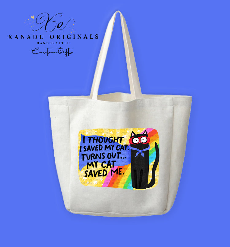 A cream-colored polyester linen tote bag featuring a rounded rectangular graphic with a textured yellow starry background and a retro rainbow arch. On the right stands a cartoon black cat wearing a red superhero mask and blue cape bow next to bold black text reading "I THOUGHT I SAVED MY CAT. TURNS OUT... MY CAT SAVED ME."