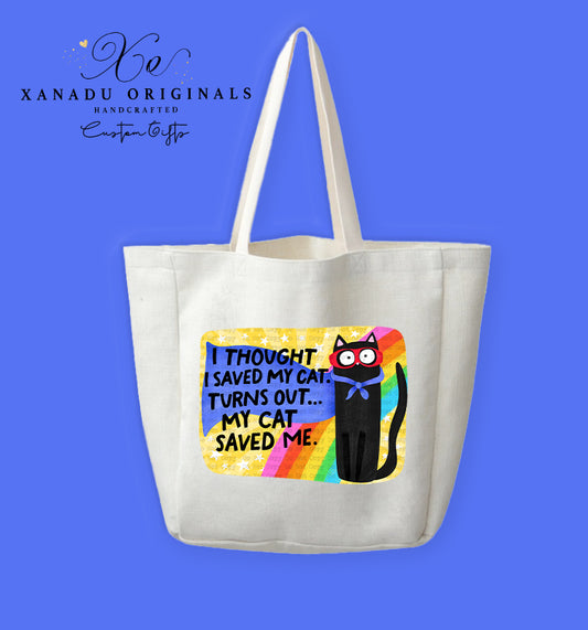 A cream-colored polyester linen tote bag featuring a rounded rectangular graphic with a textured yellow starry background and a retro rainbow arch. On the right stands a cartoon black cat wearing a red superhero mask and blue cape bow next to bold black text reading "I THOUGHT I SAVED MY CAT. TURNS OUT... MY CAT SAVED ME."