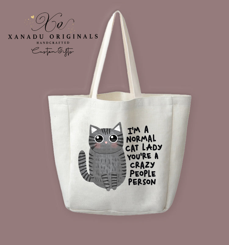 A cream-colored polyester linen tote bag featuring a cartoon illustration of a round, wide-eyed grey striped tabby cat with rosy cheeks. Bold black text to the right reads "I'M A NORMAL CAT LADY YOU'RE A CRAZY PEOPLE PERSON."