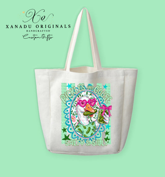 A cream-colored polyester linen tote bag featuring a bright mint and pink illustration of a goose wearing heart glasses and eating a pickle next to the text "PICKLES AND GOOSE SALTY & SILLY".