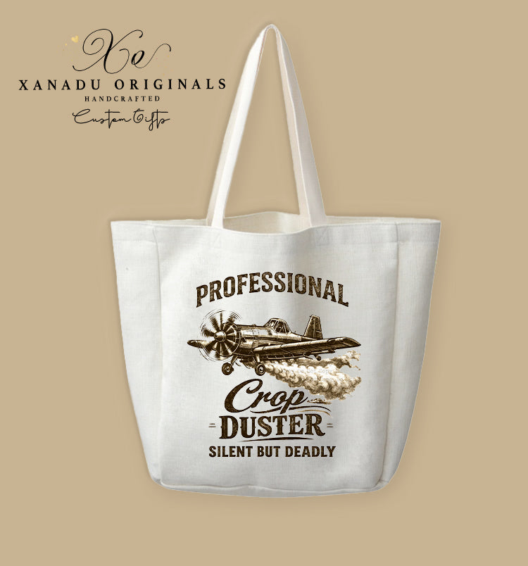 A cream-colored polyester linen tote bag featuring a sepia-toned vintage illustration of a propeller crop duster airplane in mid-flight releasing a trail of smoke. Bold black and brown block text arches above and below the graphic reading "PROFESSIONAL CROP DUSTER SILENT BUT DEADLY."