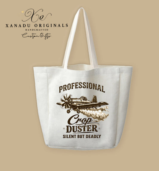 A cream-colored polyester linen tote bag featuring a sepia-toned vintage illustration of a propeller crop duster airplane in mid-flight releasing a trail of smoke. Bold black and brown block text arches above and below the graphic reading "PROFESSIONAL CROP DUSTER SILENT BUT DEADLY."