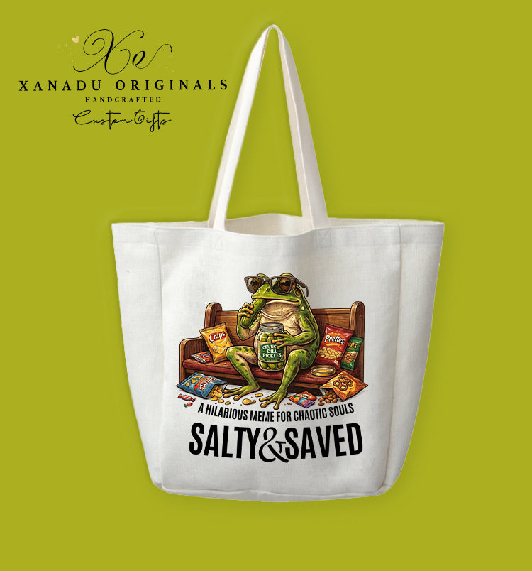 A cream-colored polyester linen tote bag featuring a colorful illustration of a green frog wearing sunglasses sitting on a wooden church pew, eating a jar of pickles surrounded by snack bags. The text reads "A HILARIOUS MEME FOR CHAOTIC SOULS" and "SALTY & SAVED" in large bold letters.