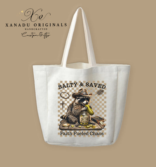 A cream-colored polyester linen tote bag featuring a rustic western illustration of a raccoon in a cowboy hat eating pickles with the text "SALTY & SAVED" and "Faith Fueled Chaos".