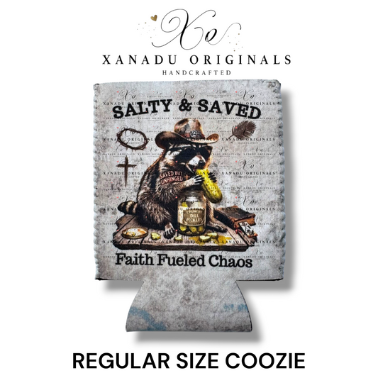 "Xanadu Originals handcrafted regular size neoprene can coozie featuring a rustic vintage illustration of a cowboy raccoon eating a dill pickle beside a cross and Bible with the text 'Salty & Saved' and 'Faith Fueled Chaos' on a gray textured background."