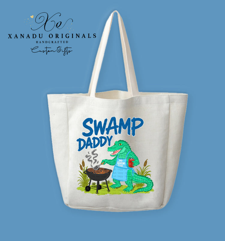 A cream-colored polyester linen tote bag featuring a cartoon illustration of a green alligator wearing a blue apron, holding a red can while flipping food on a smoking black barbecue grill in a swamp. Above the graphic, bold blue text reads "SWAMP DADDY."