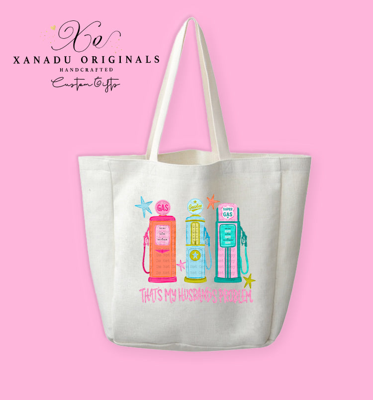 A cream-colored polyester linen tote bag featuring a retro mid-century modern graphic of three vintage gas pumps in pink, yellow, and teal with starburst details. Bright pink stylized text underneath reads "THAT'S MY HUSBAND'S PROBLEM."