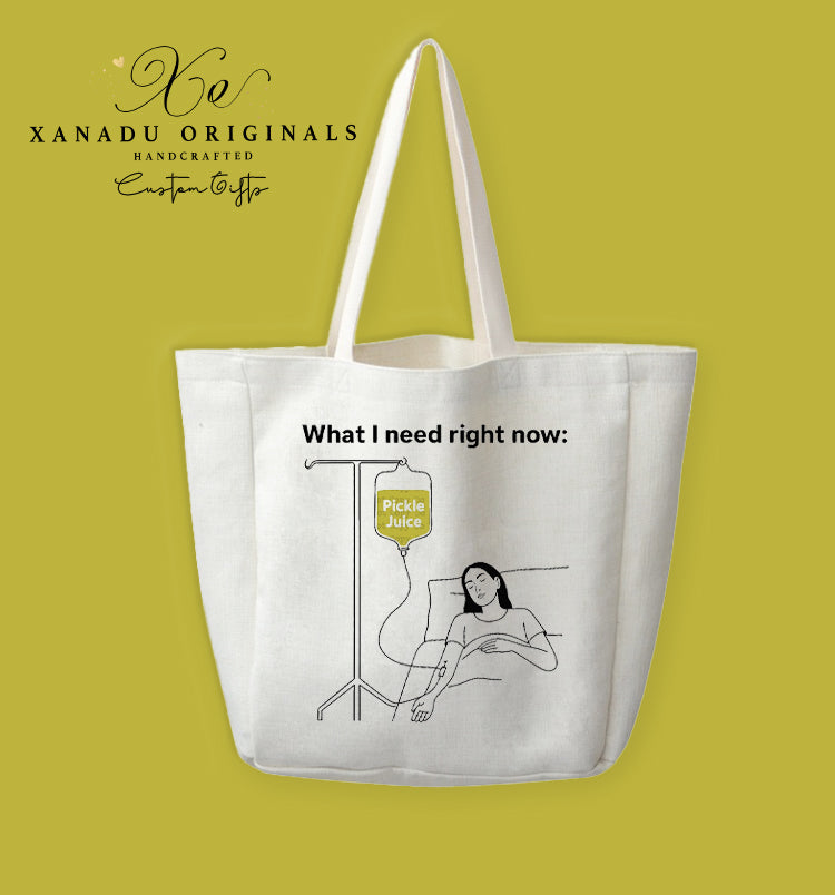 A cream-colored polyester linen tote bag featuring a minimalist black line-art illustration of a person in bed attached to an IV bag of pickle juice under the text "What I need right now:".