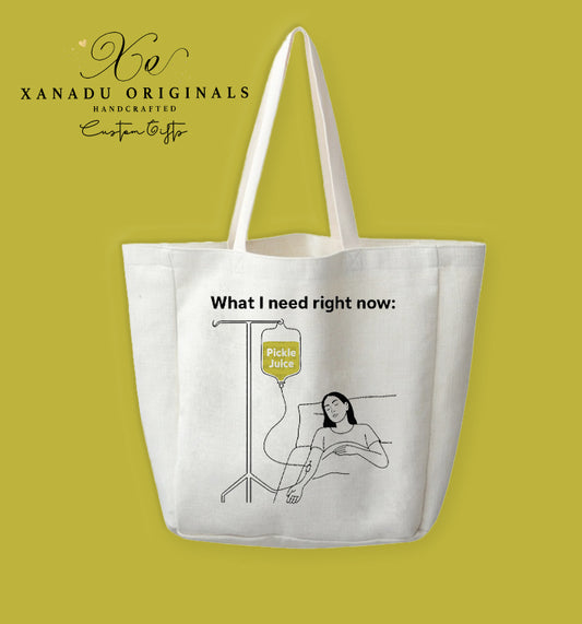 A cream-colored polyester linen tote bag featuring a minimalist black line-art illustration of a person in bed attached to an IV bag of pickle juice under the text "What I need right now:".
